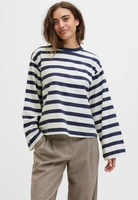 Young woman with wavy hair wearing a navy and white striped long-sleeve shirt and beige trousers, smiling confidently against a plain background.