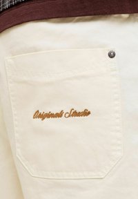 Gros plan d'une poche de pantalon crème avec le texte brodé marron « Originals Strator » et un rivet métallique dans le coin.