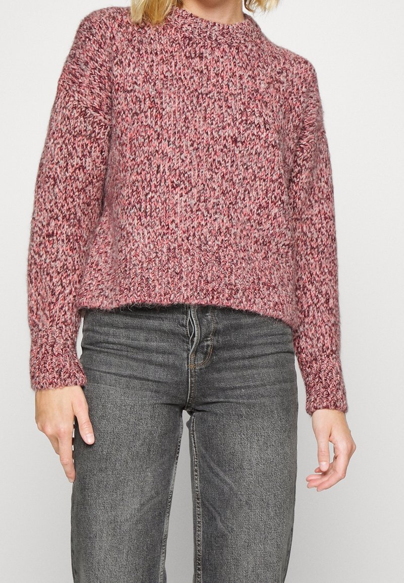 Person, die einen grob gestrickten Pullover in gemischten Rosatönen und ausgewaschenen schwarzen Jeans trägt, steht vor einem schlichten weißen Hintergrund.