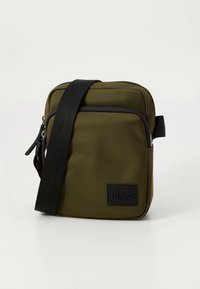 Kompakt, olívazöld válltáska fekete állítható pánttal, elülső cipzáras zsebbel és HUGO logófolttal a szöveten.