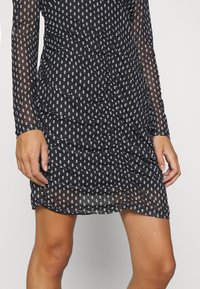 Schwarzes Kleid mit langen, durchsichtigen Ärmeln mit weißen Rautenmuster, gerafftem Design und einem gerafften Detail an der Seite.