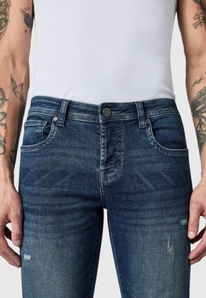 Mand iført mørkeblå slidte jeans og en hvid ribstrikket tanktop med synlige tatoveringer på begge underarme.