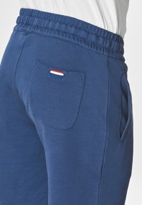 Pantaloni della tuta blu navy con cintura elastica. Presentano una tasca posteriore unica con un'etichetta tricolore. Il materiale sembra morbido con una consistenza liscia.