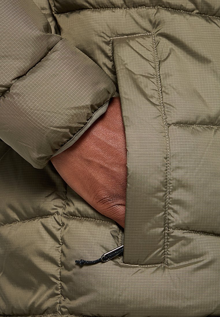 Chaqueta acolchada de color verde oliva con un exterior texturizado, bolsillo lateral con cremallera y una mano descansando dentro del bolsillo.