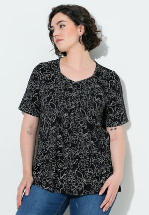 Femme aux cheveux courts et foncés portant un chemisier noir à manches courtes à motif floral et un jean bleu, debout contre un mur blanc, regardant vers la gauche.