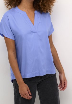 Personne aux cheveux bouclés portant un chemisier bleu clair à manches courtes et un jean noir, debout contre un mur blanc.