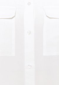 Chemise blanche boutonnée sur le devant avec deux poches poitrine et des boutons couleur perle le long de la patte centrale.