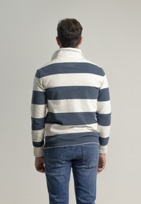 Camisa de rayas azul marino y blanca de manga larga con cuello alzado, presenta un ajuste relajado y está confeccionada en suave algodón. Lleva puesta unos vaqueros azules.