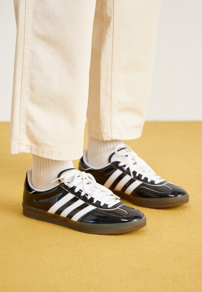 Svarta sneakers i lackläder med vita skosnören och tre vita ränder. Sulan är brun och skorna har en rundad tådesign.