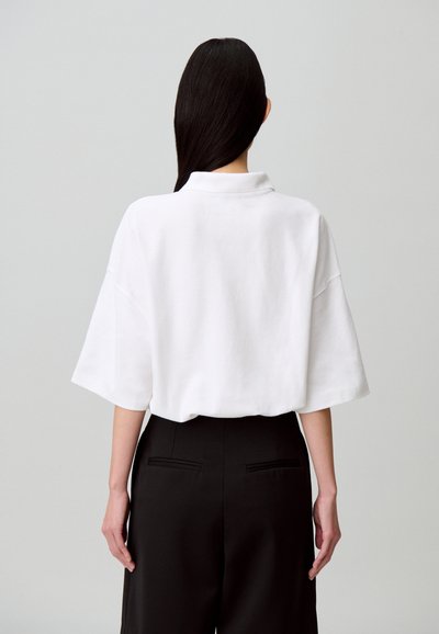 Polo blanc surdimensionné en tissu texturé, doté d'un col et de manches courtes, associé à un pantalon noir taille haute.