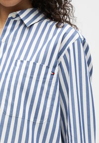 Λευκό και μπλε κατακόρυφα ριγέ πουκάμισο με κουμπιά, τσέπη στο στήθος και μικρό κεντημένο λογότυπο, που φοριέται από άτομο με σγουρά μαλλιά.