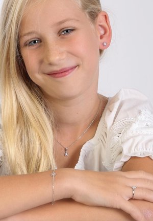 Silbernes Schmuckset mit einer Blumenanhänger-Halskette, passenden Ohrringen, einem Armband und einem Ring mit klaren Edelsteinakzenten. Weiße Bluse mit Bestickung.