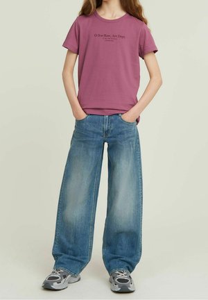 Personne portant un t-shirt graphique mauve, un jean large bleu clair et des baskets grises, debout les mains dans les poches sur un fond clair.