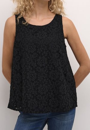 Vrouw met een mouwloze zwarte kanten top met bloemmotieven en een blauwe spijkerbroek, staand tegen een neutrale achtergrond.