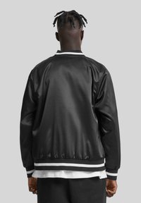 Veste bomber en satin noir avec des poignets et un ourlet rayés blancs, dotée d'un col classique et d'une coupe décontractée. Texture lisse, vue arrière montrée.