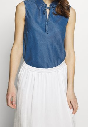 Blusa sin mangas de mezclilla en azul oscuro con cuello abotonado y cierre con lazo, combinada con una falda blanca, plisada, con cintura elástica.