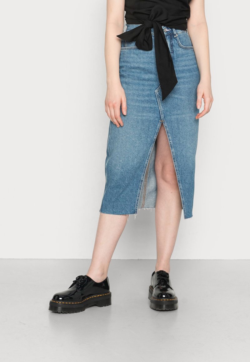 Falda lápiz de mezclilla en azul claro con una abertura frontal, dobladillo deshilachado y cintura alta. Combinada con zapatos de charol negro.