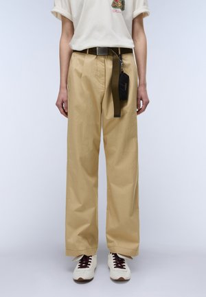 Persona che indossa pantaloni beige a gamba larga con una cintura nera e una piccola borsa nera, sneakers bianche con lacci scuri e una t-shirt bianca.