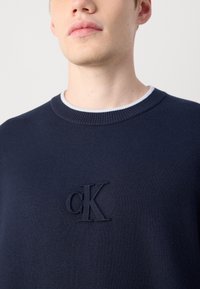 Tamsiai mėlynas megztinis, pagamintas iš minkštos mezginio medžiagos, su vertikaliu apykakle ir siuvinėtu 'cK' logotipu krūtinėje. Lygus paviršius ir paprastas dizainas.