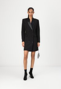 Musta kaksirivinen bleiserimekko satiinikauluksilla, jossa on rakenteelliset olkapäät, läppätaskut ja sukkela siluetti.
