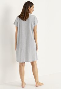 Kvinna som står barfota, vänd bort, iförd en lös kortärmad ljusgrå knälång klänning mot en enkel vit bakgrund.