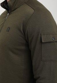 Vihreä vetoketjullinen fleece, jossa on texturoitu pinta, rintalogolla ja läpällä vasemmassa hihaosassa. Rakenne hartioille.