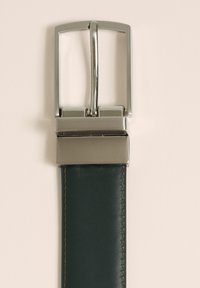 Zaļš ādas josts ar sudraba taisnstūrveida sprādzi, kurai ir pulēta apdare un izturīga eņģe, papildināta ar kontrastējošām dzeltenām šuvēm.