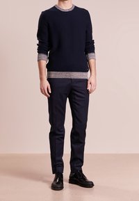 Homme portant un pull bleu marine avec des poignets et un ourlet gris, un pantalon sombre ajusté et des chaussures en cuir noir, debout devant un fond beige.