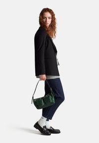 Zelená saténová kabelka přes rameno s dvěma předními kapsami, kterou drží osoba v černém saku, šedém svetru a tmavých džínách. Viditelné černé polobotky a bílé ponožky.