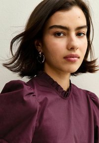 Bordeauxblus med puffärmar och volangkrage; modellen bär stora hjärtformade örhängen, håret hänger löst runt ansiktet, enkel bakgrund.