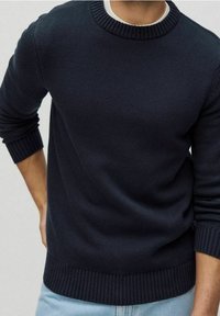 Uomo che indossa un maglione lavorato a maglia di un blu navy scuro con polsini, colletto e orlo a coste, abbinato a jeans azzurri.