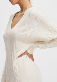 Cremiger, kabelgestrickter Pullover mit V-Ausschnitt, weiten Ärmeln, strukturiertem Muster an den Seiten und gerippten Details an den Bündchen und dem Saum.