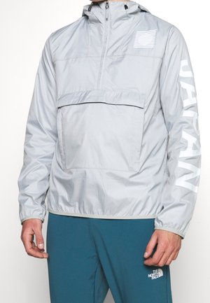 Homme portant un coupe-vent gris clair avec une poche avant et le texte "Urban" sur la manche, associé à un pantalon bleu The North Face.