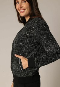 Zwart bomberjack met een ronddraaiend wit patroon, ritssluiting en geribde manchetten en zoom. Gemaakt van een gestructureerde stof.