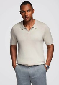 Camiseta polo de manga corta beige con cuello, hecha de tela texturizada. Combinada con pantalones de rayas gris claro y un reloj plateado.
