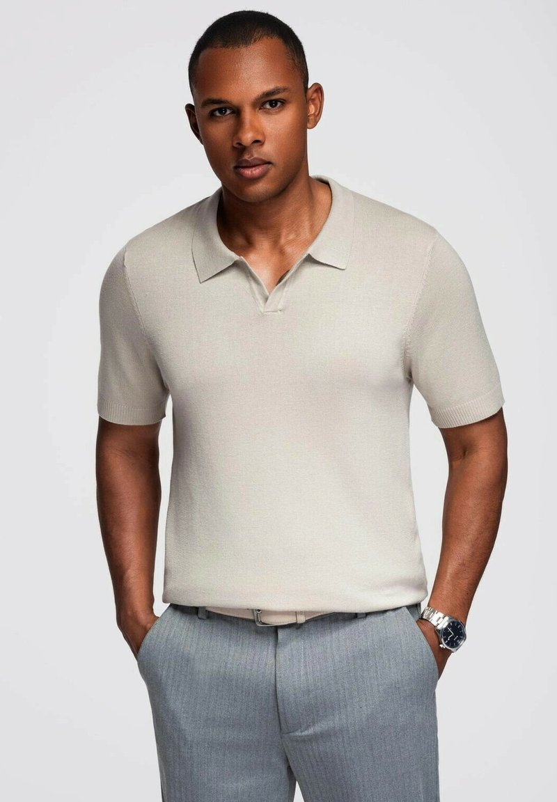 Camiseta polo de manga corta beige con cuello, hecha de tela texturizada. Combinada con pantalones de rayas gris claro y un reloj plateado.