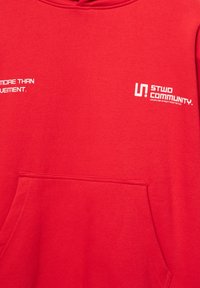 Punainen huppari, jossa on kengurutasku ja valkoinen painettu teksti "STWD COMMUNITY" sekä "MORE THAN MOVEMENT." Pehmeä kangaspinta.