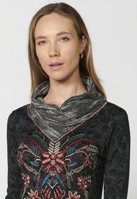 Schwarzes Oberteil mit aufwendiger, farbenfroher Blumenstickerei, kombiniert mit einem strukturierten, gefalteten grauen Schal. Glatte Materialien mit unterschiedlichen Mustern.