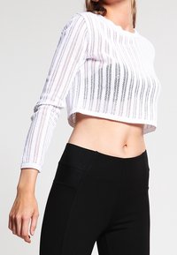 Haut blanc court à manches longues en tissu transparent avec des découpes rayées verticales. Associé à un pantalon noir taille haute avec des accents texturés.