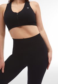 Reggiseno sportivo nero con zip anteriore, tessuto testurizzato e dettaglio con logo. Abbinato a leggings neri a vita alta, entrambi realizzati in materiale elastico.
