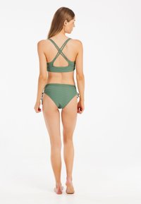 Žalias bikini komplektas su kryžminėmis juostelėmis, tekstūruota medžiaga ir raišteliais ant šonų. Dizainas pabrėžia komfortą ir palaikymą.