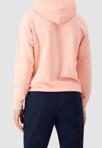 Felpa rosa chiaro con cappuccio rotondo, realizzata in tessuto morbido, indossata sopra pantaloni blu scuro. Il design presenta polsini e orlo a coste.