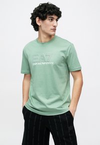 Világoszöld rövid ujjú pamut póló, fehér "EA7 Emporio Armani" logóval, stilizált betűtípussal a póló elején.
