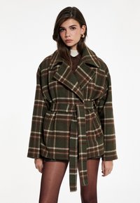 Abrigo a cuadros verdes y marrones con cuello ancho, cintura ceñida con cinturón y bolsillos laterales. Tejido suave con un ajuste relajado y mangas anchas.