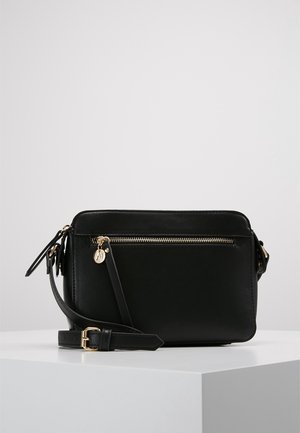 Zwarte leren schoudertas met gouden ritssluiting en gesp, verstelbare riem en een klein rond hangertje aan de ritstrekker, tentoongesteld op een wit voetstuk.