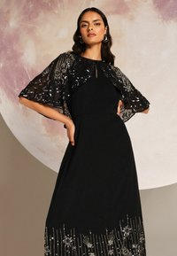 Fekete ruha gyöngyökkel díszített, hímzett fekete köpenyével, amelynek szegélyén virágos és vonalas minták találhatók. Sima anyagból készült, testhez álló szabású.