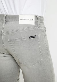 Šedé džíny z denimu mají jednoduchou zadní kapsu s malou černou štítkem "CALVIN KLEIN JEANS" a bílou značkovou štítkem na pase.