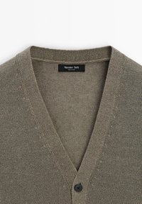 Grauer Strickcardigan mit V-Ausschnitt, ausgestattet mit einer schwarzen Knopfschließe und einem Etikett. Die Textur ist glatt und leicht.