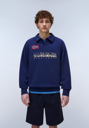 Jeune homme aux cheveux bouclés portant un sweat-shirt Napapijri bleu marine avec un écusson du drapeau norvégien et un short noir, debout devant un fond uni.
