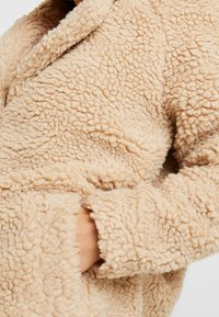 Béžový kožich z umělé kožešiny s texturovaným, plyšovým povrchem, se šálovým límcem a bočními kapsami. Měkký, pohodlný materiál s uvolněným střihem.